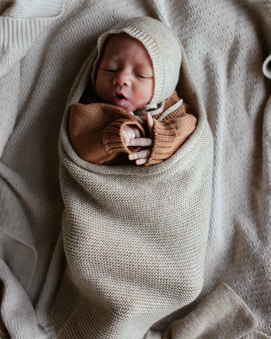 Baby med strikhue i Hvid merinould cocoon Sand på et strikket tæppe