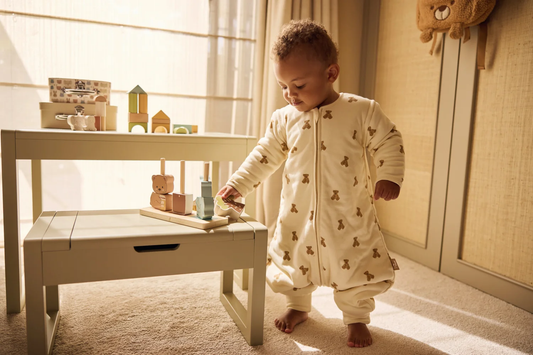 Barn i Jollein Teddy sovepose med bamseprint står ved legetøj med bare fødder