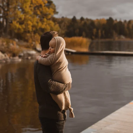 Baby i merinould cocoon oat fra Hvid, holdt i armene udendørs ved sø – giver tryghed, varme og komfort i kølige måneder.