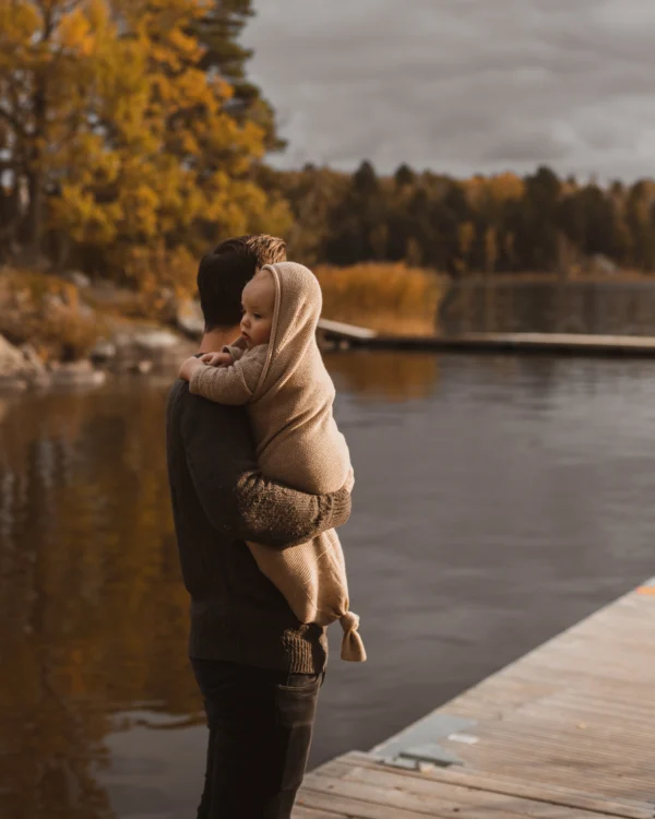 Baby i merinould cocoon oat fra Hvid, holdt i armene udendørs ved sø – giver tryghed, varme og komfort i kølige måneder.